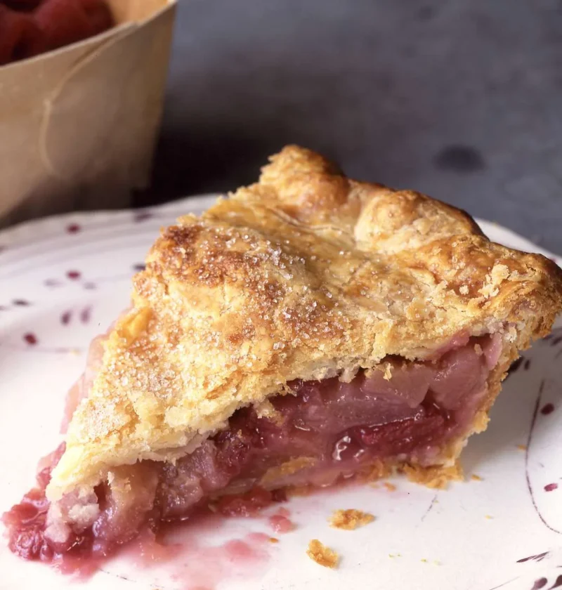 Slice of Martha Stewart apple-raspberry pie with a golden sugar-sprinkled crust and a juicy pink fruit filling on a speckled plate