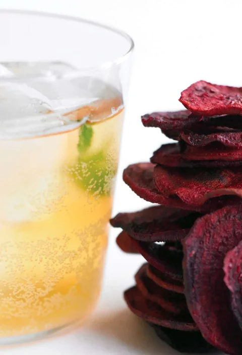 Martha Stewart apple spritzers in a glass with ice and a mint sprig next to a stack of crisp beet chips