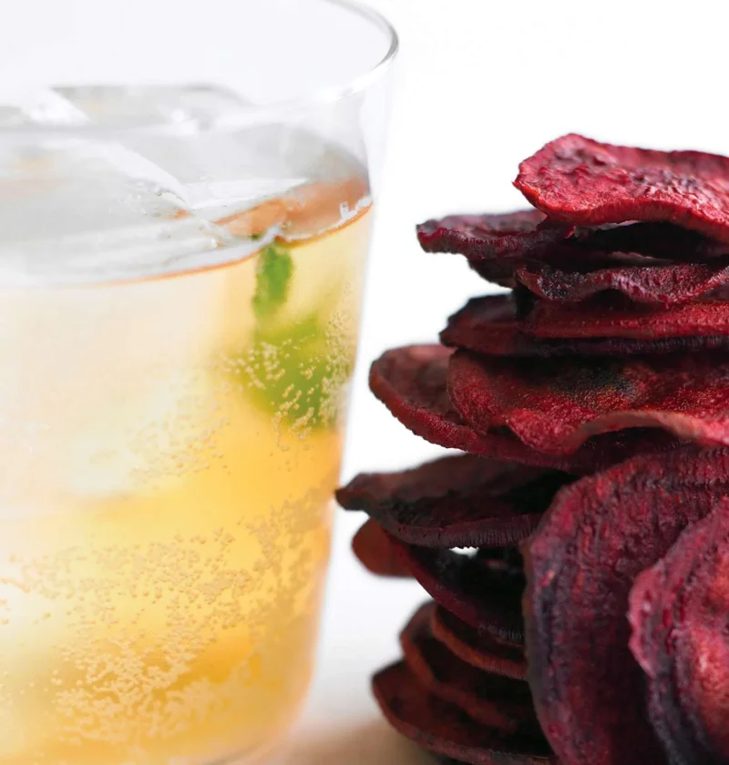 Martha Stewart apple spritzers in a glass with ice and a mint sprig next to a stack of crisp beet chips