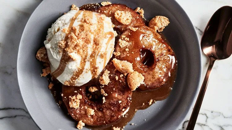 Martha Stewart apple sundaes with cider caramel sauce, baked apple rounds topped with vanilla ice cream, caramel and crushed cookies in a gray bowl