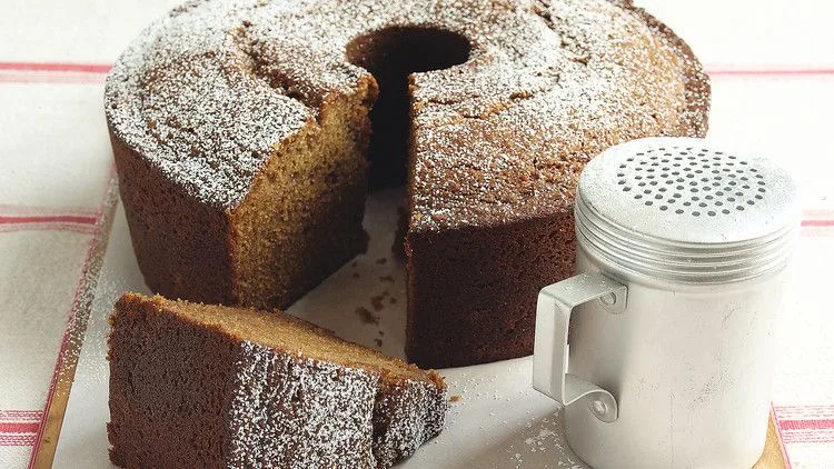 Martha Stewart applesauce cake dusted with confectioners' sugar on a board with a slice cut and a sugar shaker beside it