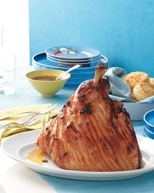 Martha Stewart apricot-bourbon glazed ham, scored and golden brown, on a white platter with a bowl of glaze and biscuits