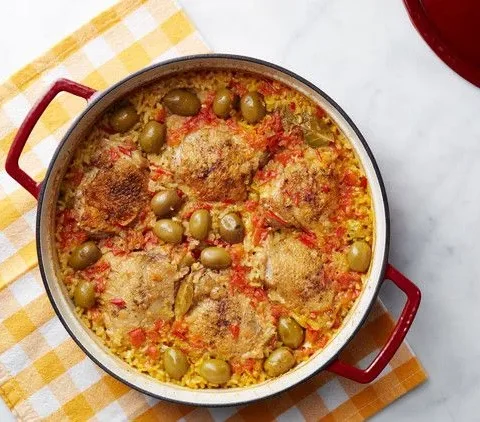 Martha Stewart arroz con pollo in a red Dutch oven with browned chicken thighs, green olives and saffron rice on a yellow checked towel