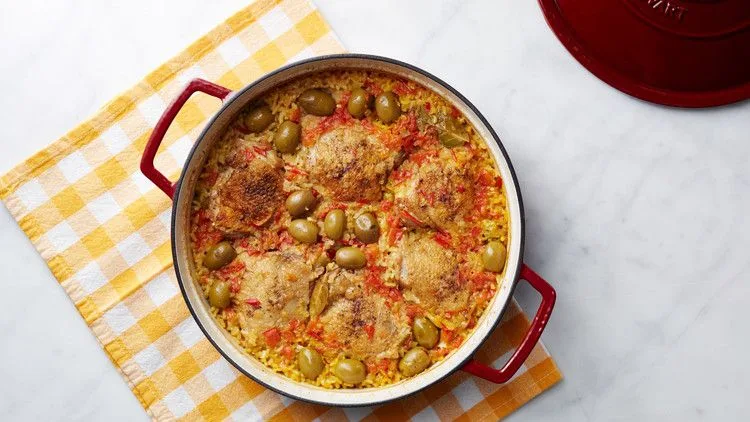 Martha Stewart arroz con pollo in a red Dutch oven with browned chicken thighs, green olives and saffron rice on a yellow checked towel