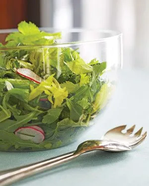 Martha Stewart arugula salad with radishes and caper vinaigrette in a clear glass bowl with serving utensils