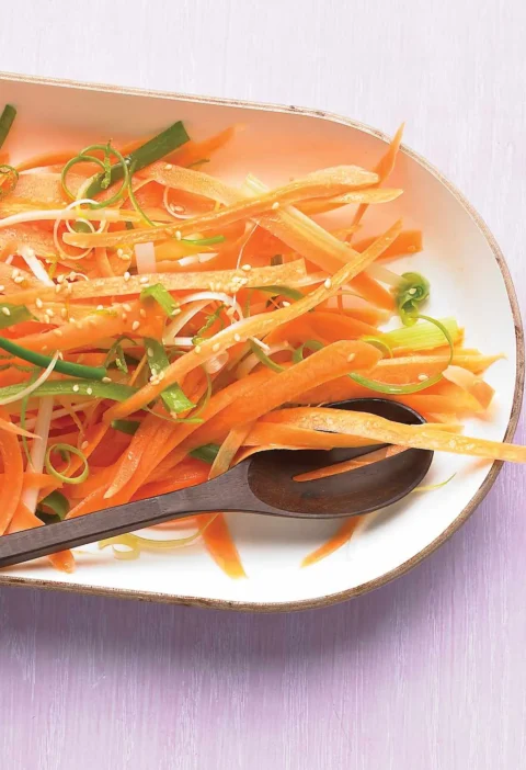 Martha Stewart Asian carrot slaw with carrot ribbons, sliced scallions and sesame seeds on an oval white platter with a wooden spoon