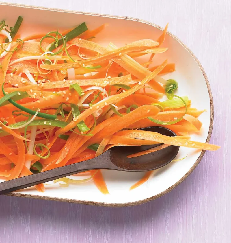 Martha Stewart Asian carrot slaw with carrot ribbons, sliced scallions and sesame seeds on an oval white platter with a wooden spoon