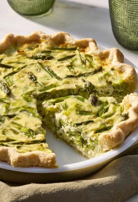 Martha Stewart asparagus, leek and Gruyere quiche with one slice cut out, on a white pie plate with plates and gold forks nearby
