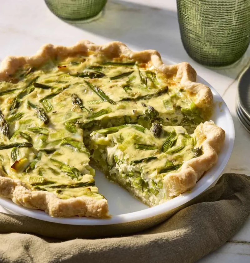 Martha Stewart asparagus, leek and Gruyere quiche with one slice cut out, on a white pie plate with plates and gold forks nearby