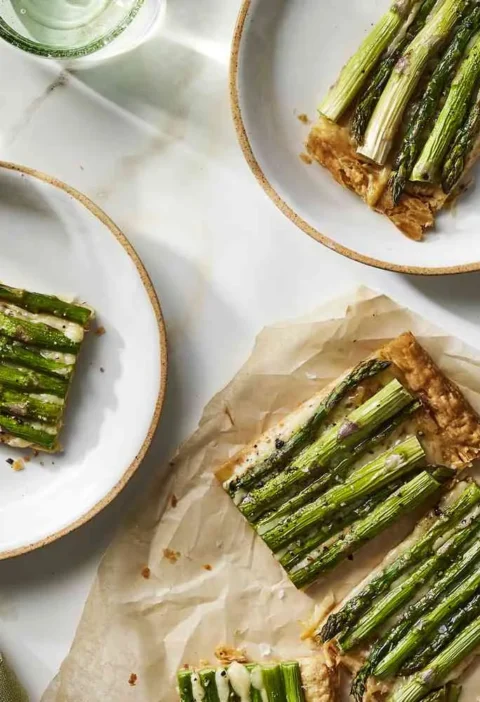 Martha Stewart asparagus tart with Gruyère cut into slices on plates and parchment, topped with rows of roasted asparagus spears