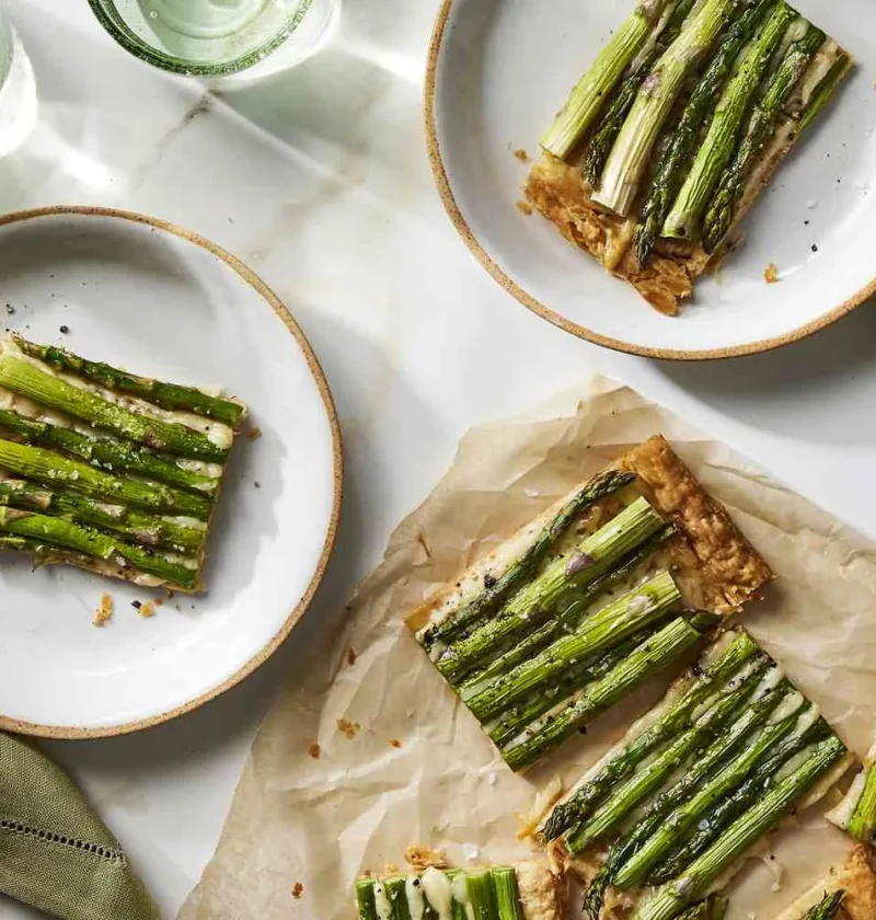 Martha Stewart asparagus tart with Gruyère cut into slices on plates and parchment, topped with rows of roasted asparagus spears