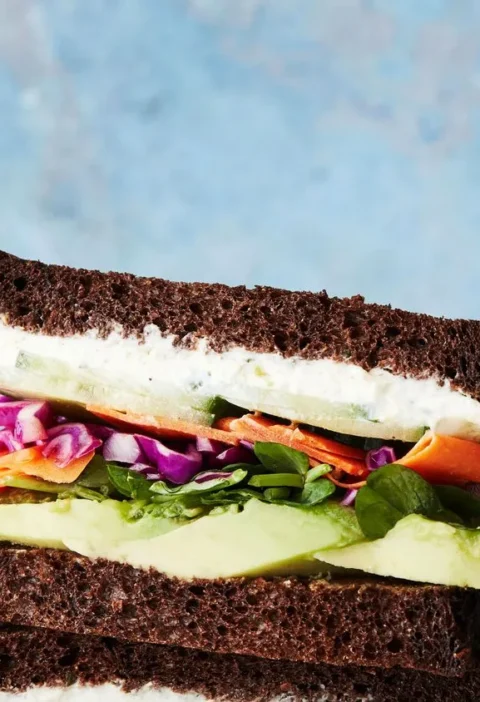 Martha Stewart avocado-and-Boursin sandwich on pumpernickel with red cabbage, carrot ribbons and sliced avocado stacked against a blue background