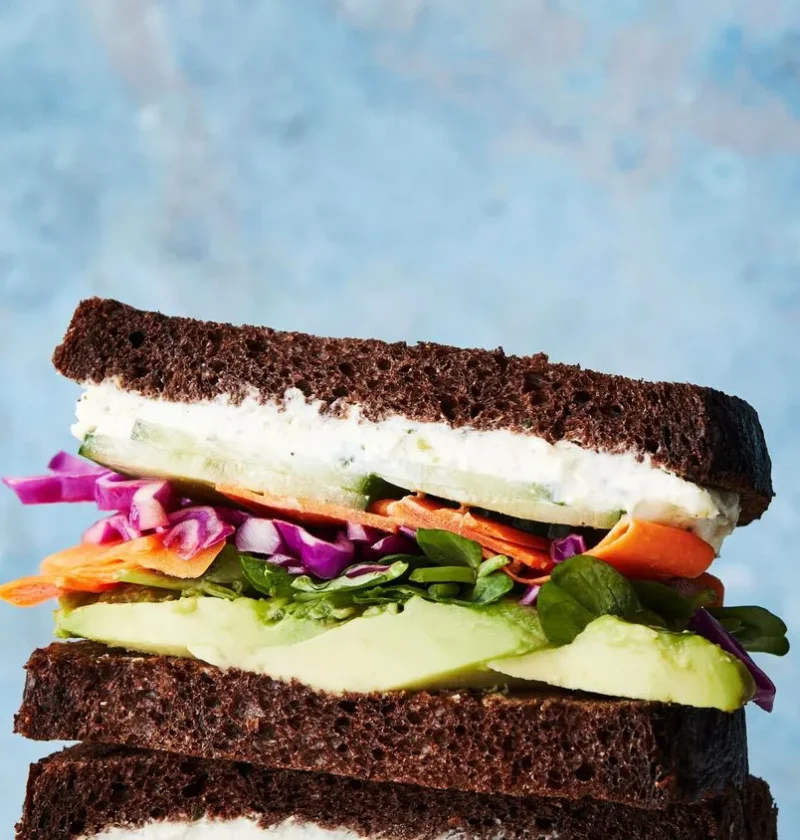 Martha Stewart avocado-and-Boursin sandwich on pumpernickel with red cabbage, carrot ribbons and sliced avocado stacked against a blue background