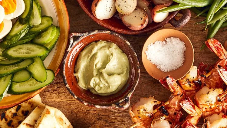 Martha Stewart avocado cream sauce in a rustic clay bowl next to grilled shrimp, cucumber slices, potatoes and green beans