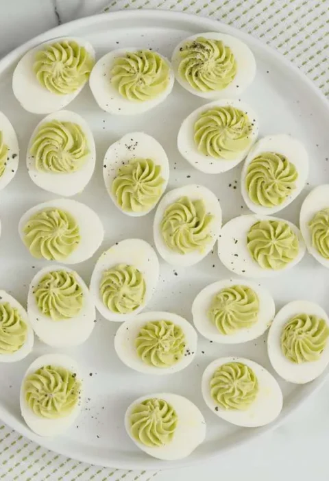 Martha Stewart avocado deviled eggs with pale green piped ruffled filling and black pepper on a round white platter