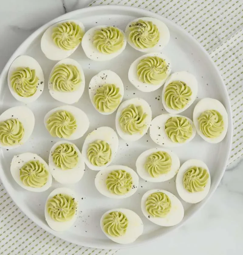 Martha Stewart avocado deviled eggs with pale green piped ruffled filling and black pepper on a round white platter