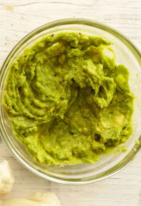 Martha Stewart avocado mash in a small glass bowl on a white wooden surface with cauliflower florets nearby