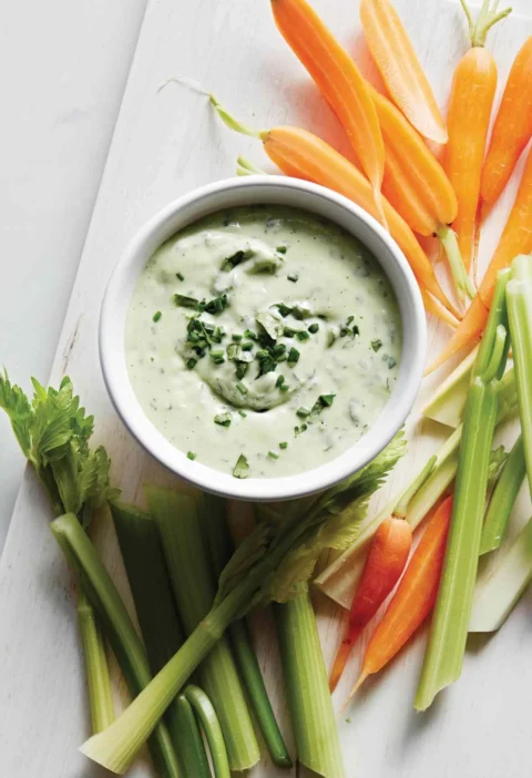Martha Stewart avocado ranch dip in a white bowl topped with chopped herbs, surrounded by baby carrots and celery sticks