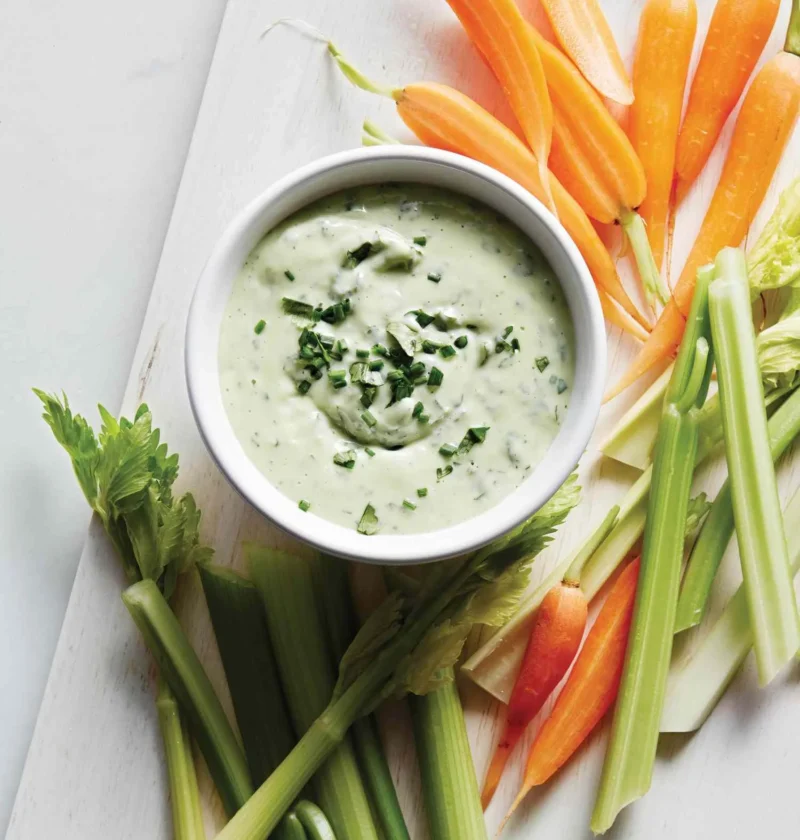Martha Stewart avocado ranch dip in a white bowl topped with chopped herbs, surrounded by baby carrots and celery sticks