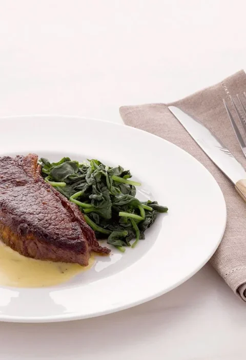 Martha Stewart Béarnaise sauce pooled on a white plate beside a seared steak and sautéed greens