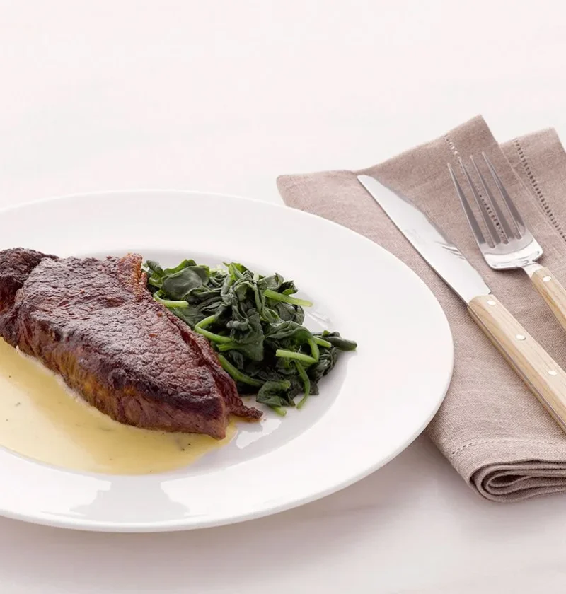 Martha Stewart Béarnaise sauce pooled on a white plate beside a seared steak and sautéed greens
