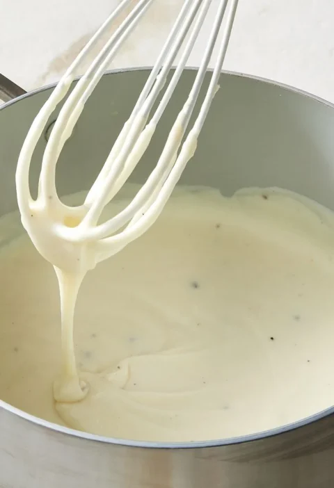 Martha Stewart béchamel sauce for lasagna in a saucepan with a whisk lifting a ribbon of creamy white sauce