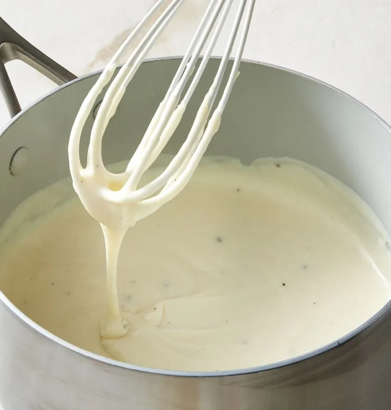 Martha Stewart béchamel sauce for lasagna in a saucepan with a whisk lifting a ribbon of creamy white sauce