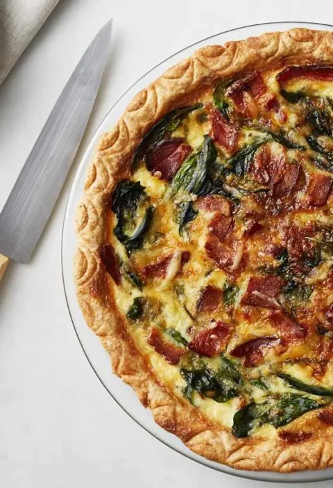 Martha Stewart bacon and spinach quiche in a glass pie plate with a golden crimped crust, crisp bacon and wilted spinach on top