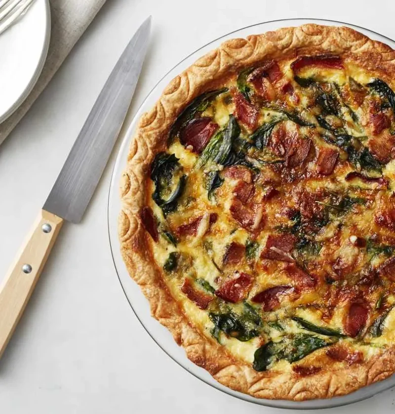 Martha Stewart bacon and spinach quiche in a glass pie plate with a golden crimped crust, crisp bacon and wilted spinach on top