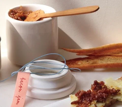 Martha Stewart bacon jam in a white jar with a wooden spoon beside toasted baguette slices and a crostini topped with bacon jam and cheese