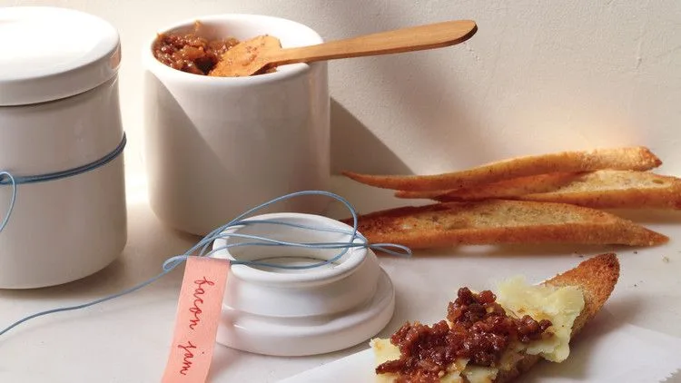 Martha Stewart bacon jam in a white jar with a wooden spoon beside toasted baguette slices and a crostini topped with bacon jam and cheese