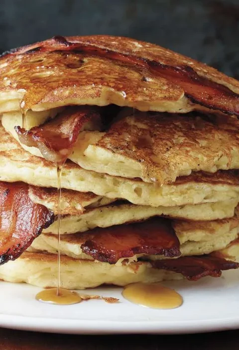 Martha Stewart bacon pancakes stacked on a white plate with crisp bacon slices and maple syrup dripping down