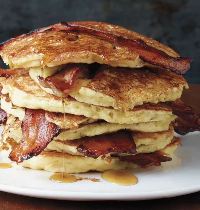 Martha Stewart bacon pancakes stacked on a white plate with crisp bacon slices and maple syrup dripping down