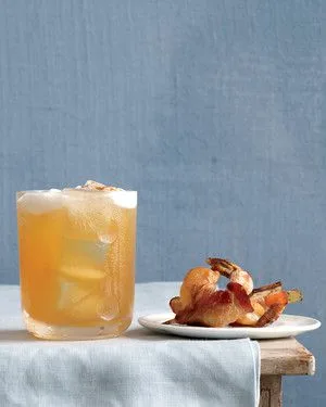 Martha Stewart bacon-wrapped ginger shrimp with crisp bacon on a white plate next to a glass of iced amber drink