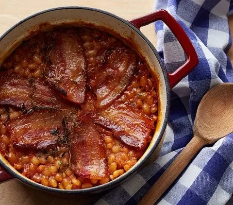 Martha Stewart baked beans with bacon in a red Dutch oven topped with browned bacon strips and thyme
