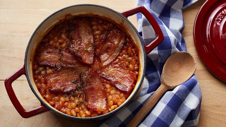 Martha Stewart baked beans with bacon in a red Dutch oven topped with browned bacon strips and thyme