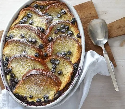 Martha Stewart baked blueberry French toast casserole with golden brioche slices and blueberries dusted with confectioners' sugar in an oval baking dish