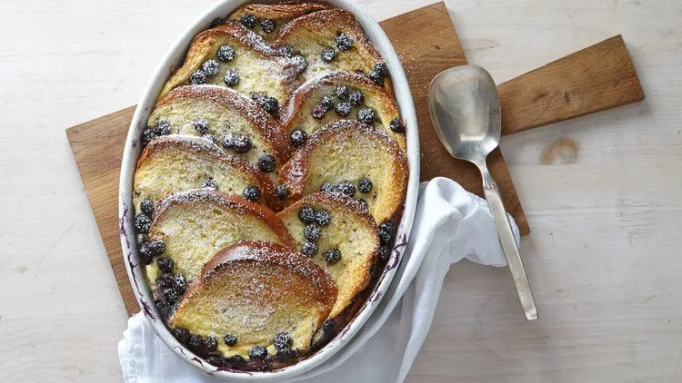 Martha Stewart baked blueberry French toast casserole with golden brioche slices and blueberries dusted with confectioners' sugar in an oval baking dish