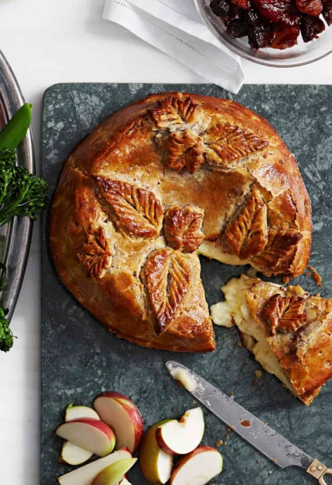 Martha Stewart baked brie with boozy fruit in golden puff pastry decorated with leaf cutouts on a marble board with apple wedges