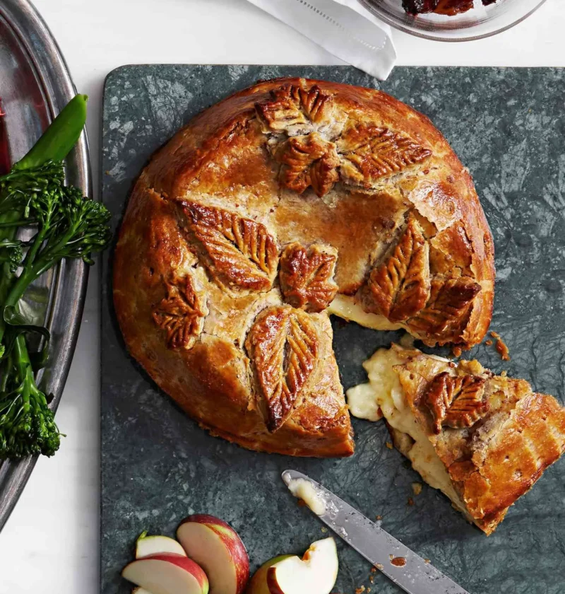 Martha Stewart baked brie with boozy fruit in golden puff pastry decorated with leaf cutouts on a marble board with apple wedges