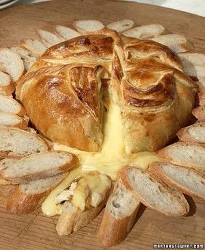Martha Stewart baked brie in golden puff pastry with melted cheese oozing out, surrounded by sliced baguette on a wooden board