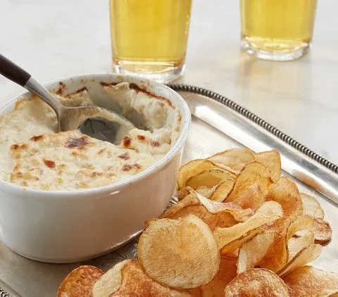 Martha Stewart baked clam dip in a white dish with a spoon, served with homemade potato chips and two glasses of beer