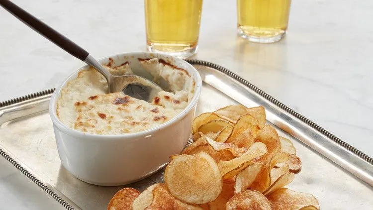 Martha Stewart baked clam dip in a white dish with a spoon, served with homemade potato chips and two glasses of beer
