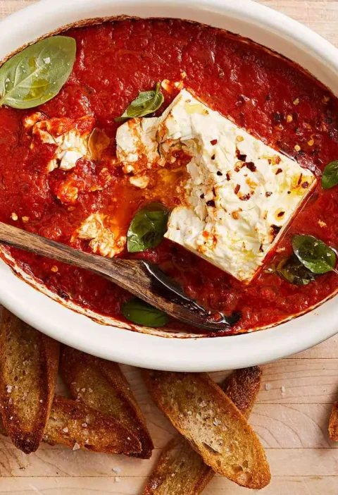 Martha Stewart baked feta-marinara dip in an oval dish with basil leaves and red-pepper flakes, served with crostini on a wooden board