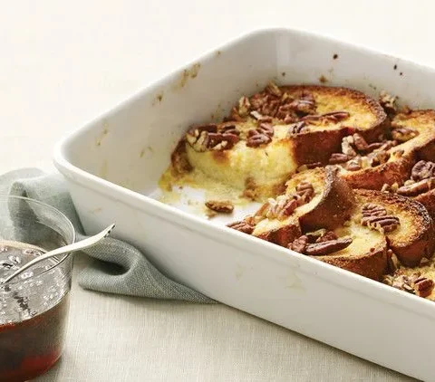 Martha Stewart baked French toast with golden brioche and pecans in a white baking dish next to a glass of rum-raisin maple syrup