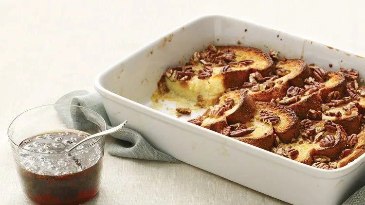 Martha Stewart baked French toast with golden brioche and pecans in a white baking dish next to a glass of rum-raisin maple syrup