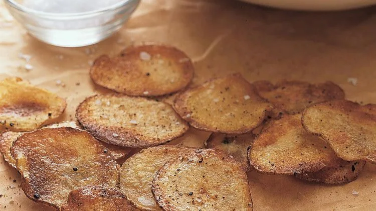 Martha Stewart baked potato chips, golden and crisp, scattered on parchment paper next to a small bowl of coarse salt