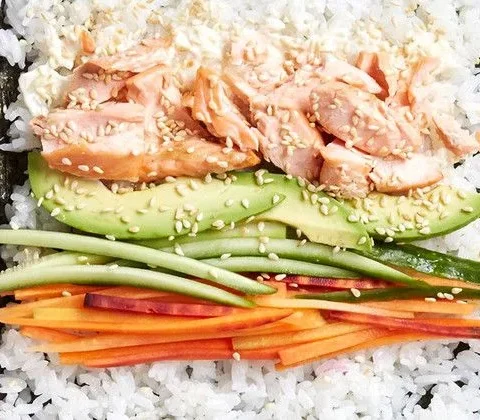 Martha Stewart baked salmon sushi rolls laid out on nori with rice, flaked salmon, avocado, cucumber and carrot on a bamboo mat