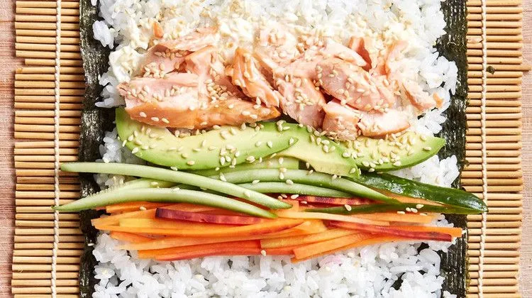 Martha Stewart baked salmon sushi rolls laid out on nori with rice, flaked salmon, avocado, cucumber and carrot on a bamboo mat
