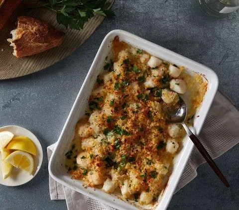 Martha Stewart baked scallops in a white baking dish topped with golden panko and parsley, next to lemon wedges and crusty bread